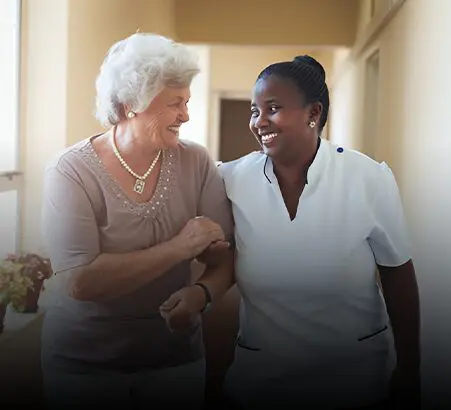 An elderly caucasian woman laughing while walking with support from a cheerful african descent nurse in a sunny corridor.