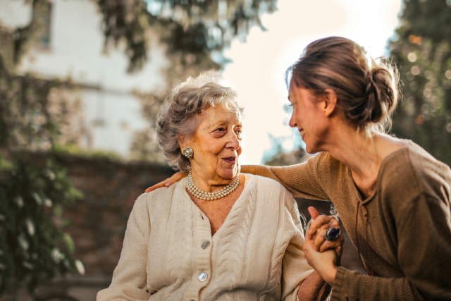 An elderly woman in a white cardigan smiles while talking with a younger woman in a brown sweater outdoors.