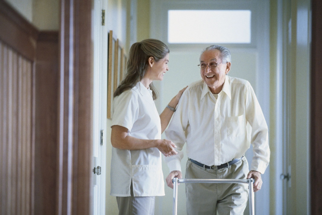 A nurse assisting an elderly man using a walker in a hallway, both smiling and engaging in conversation.
