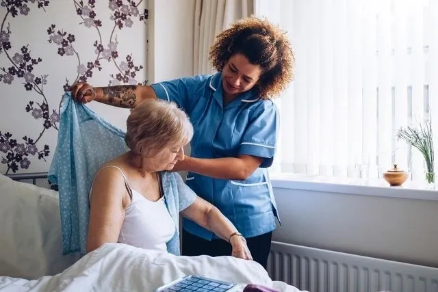 A nurse helps an elderly woman put on a blue shirt in a bright room, indicating care and assistance.